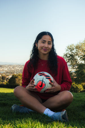 Female soccer player sitting in the grass with a soccer ball in her lap.